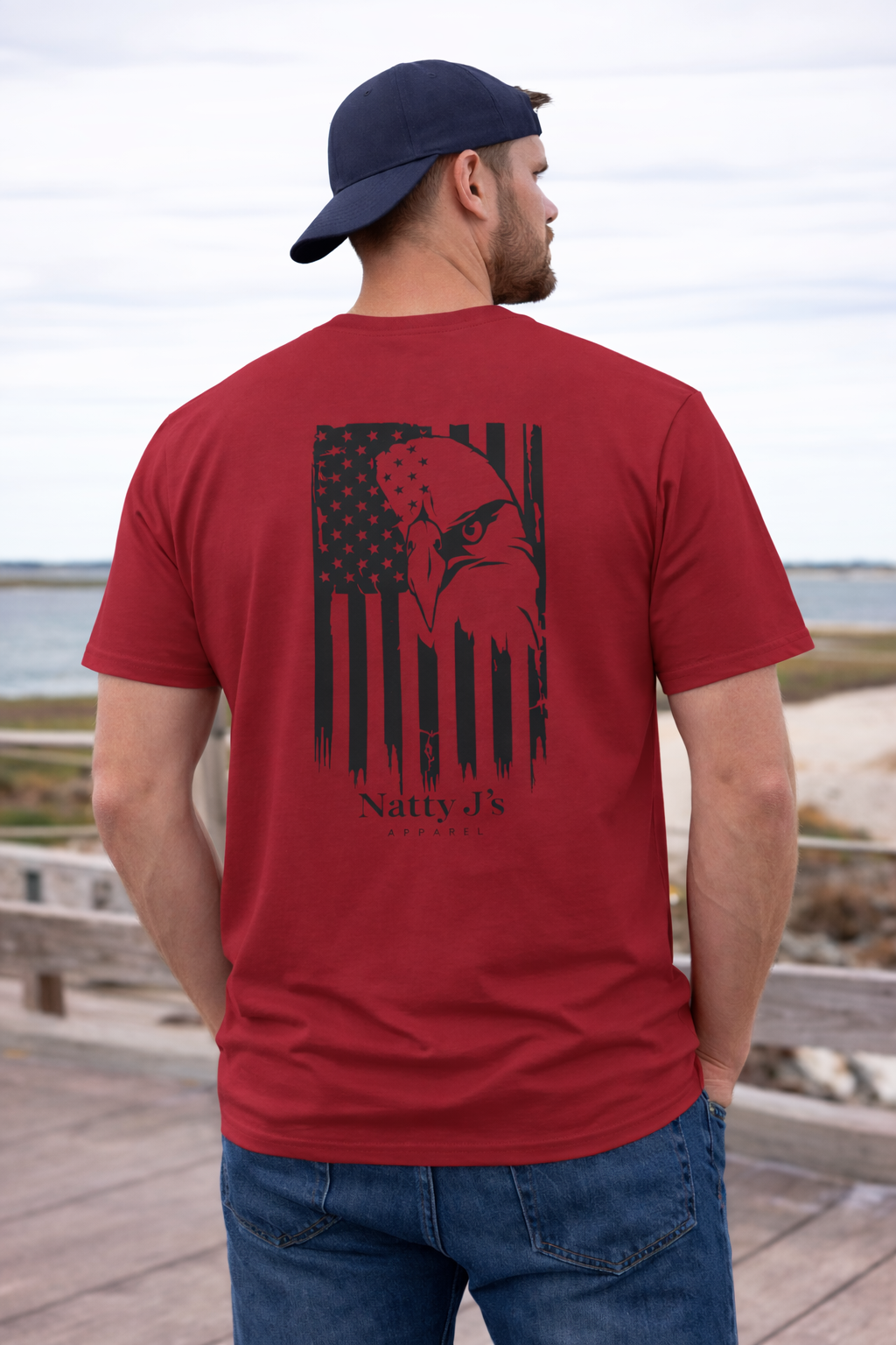 Man wearing a red t-shirt with a graphic design and text, standing on a wooden deck by a body of water.