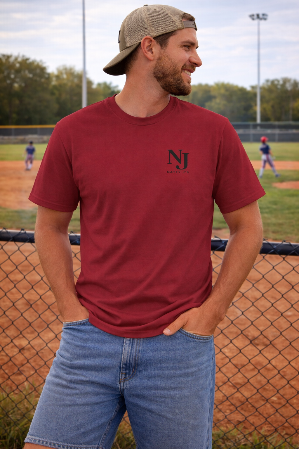 Man wearing a red t-shirt with a logo and blue jeans standing on a baseball field.
