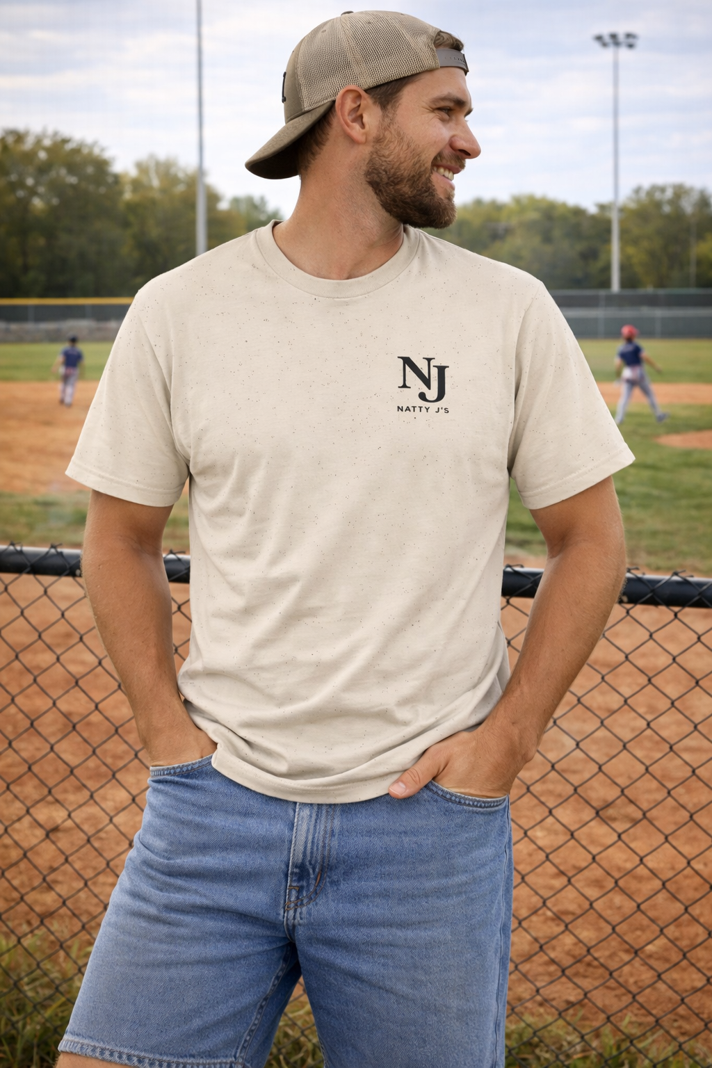 Man wearing a beige t-shirt with 'NJ' logo at a baseball field