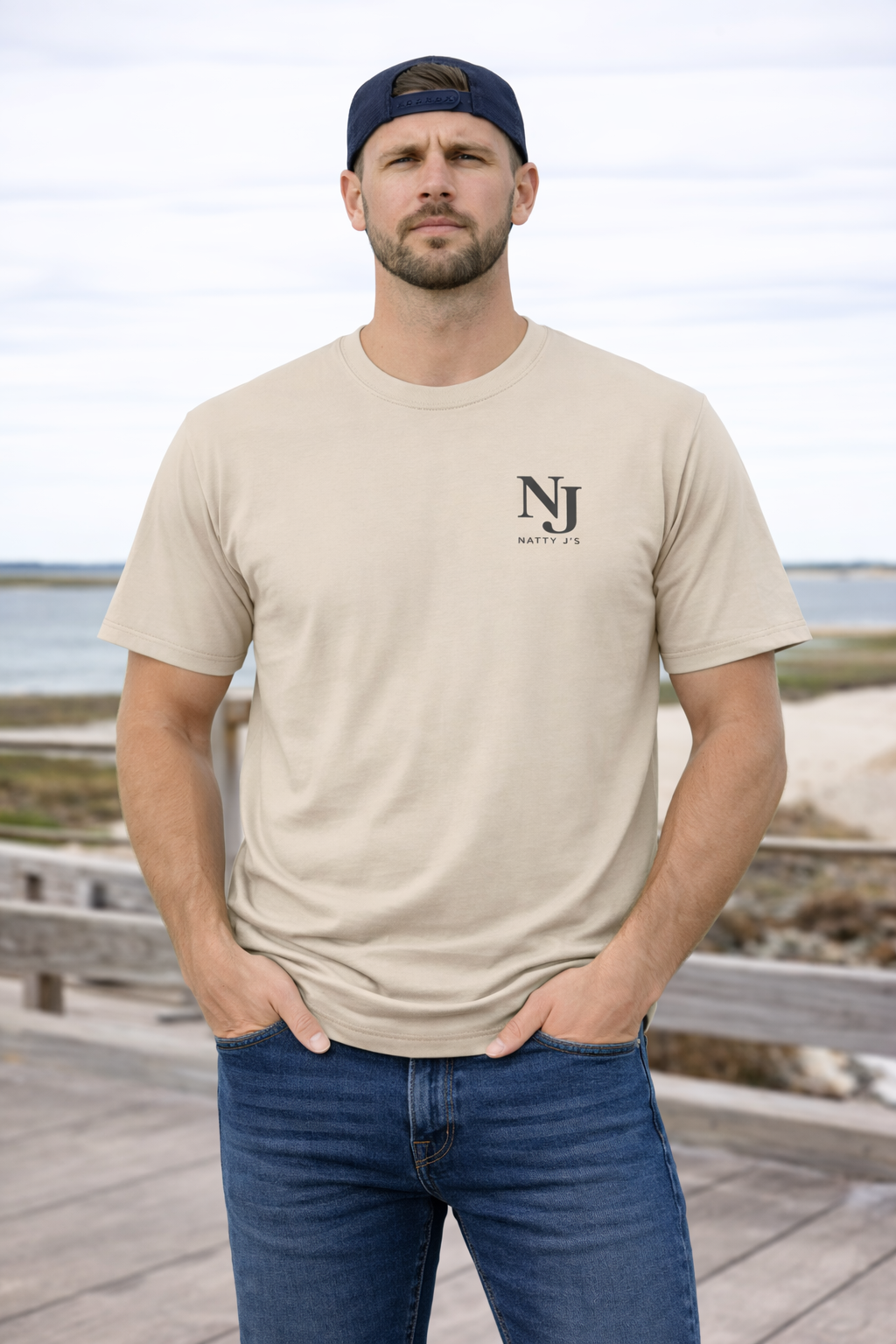 Man wearing a beige t-shirt with a logo and blue jeans standing on a wooden boardwalk by the beach.