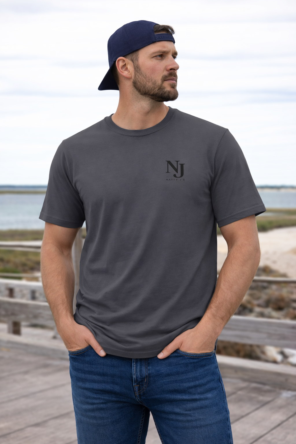 Man wearing a gray t-shirt with a logo and blue jeans standing on a wooden boardwalk by a body of water.