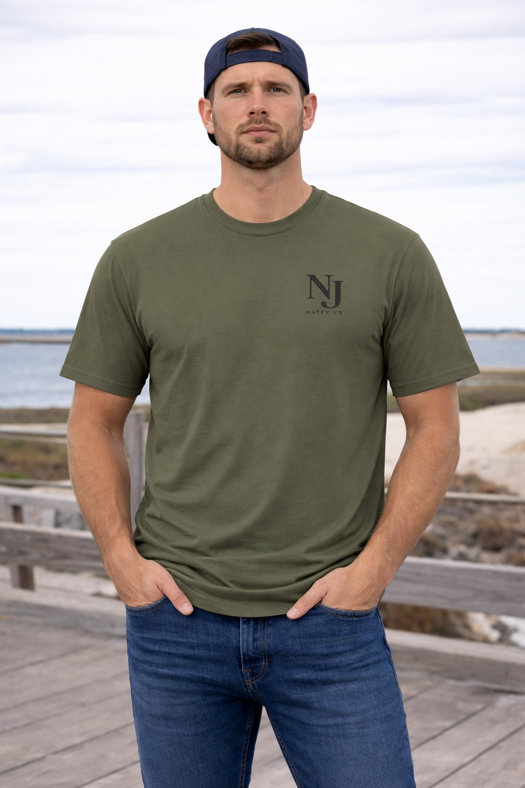Man wearing an olive green t-shirt with a logo, standing on a wooden boardwalk by the beach.