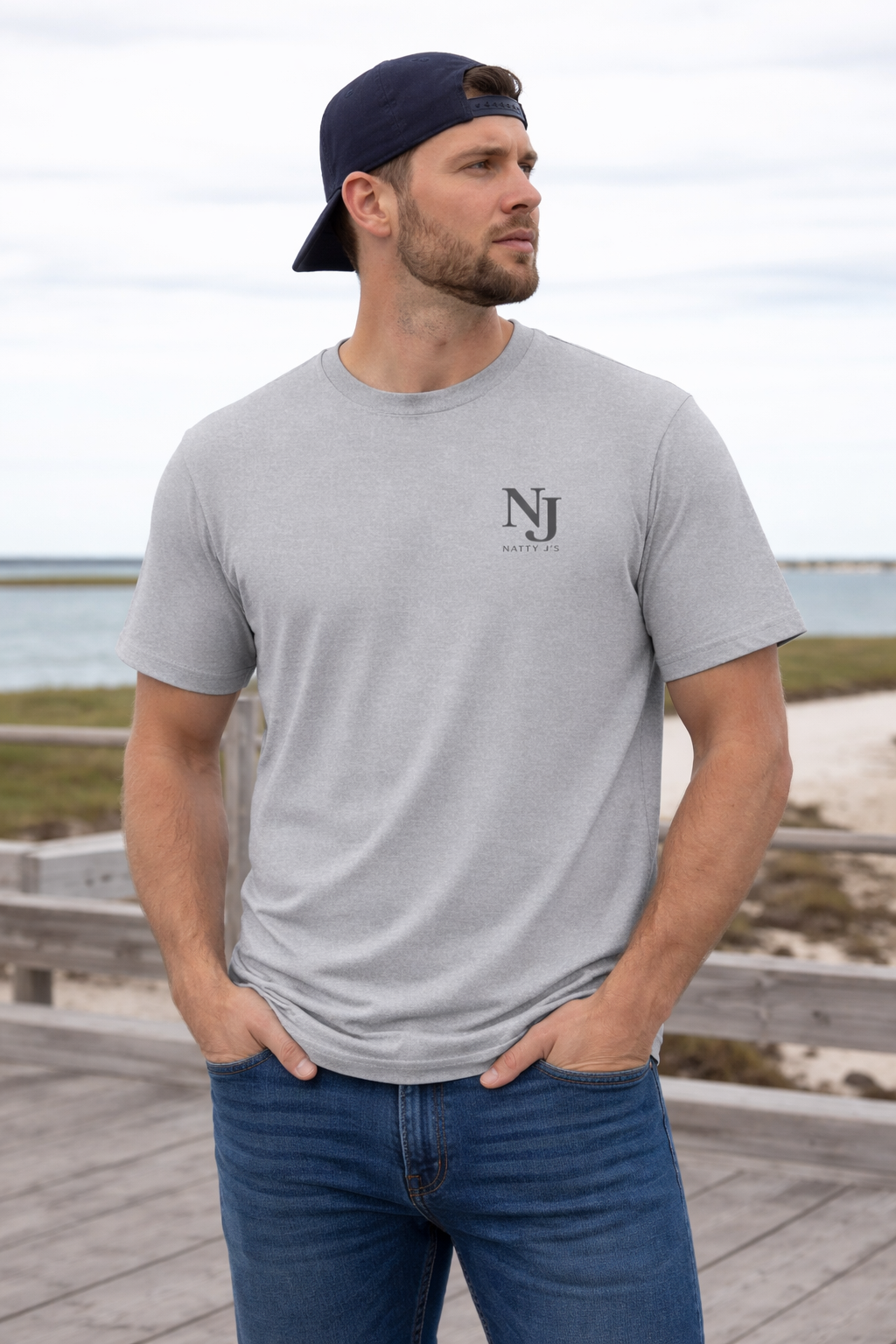 Man wearing a gray t-shirt with a logo and blue jeans standing on a wooden boardwalk by the beach.