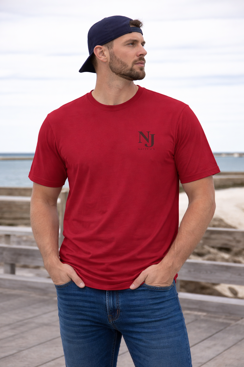Man wearing a red t-shirt with a logo and blue jeans standing on a wooden boardwalk by the beach.