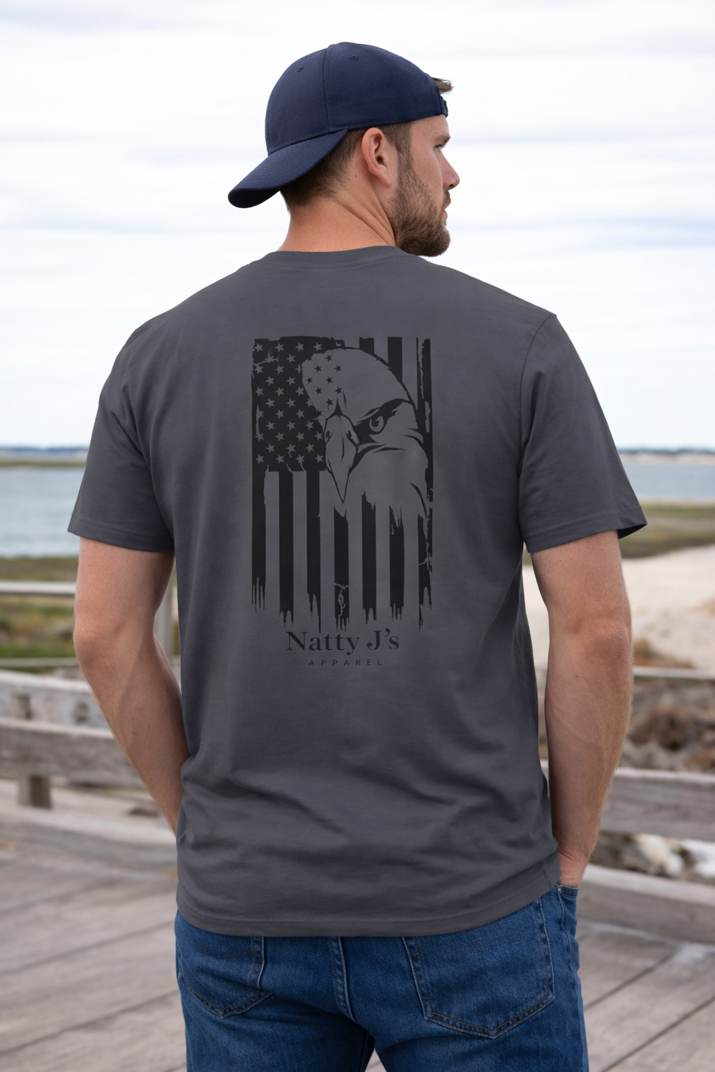 Man wearing a gray t-shirt with an eagle design and 'Natty J's' branding, standing on a wooden dock by water.