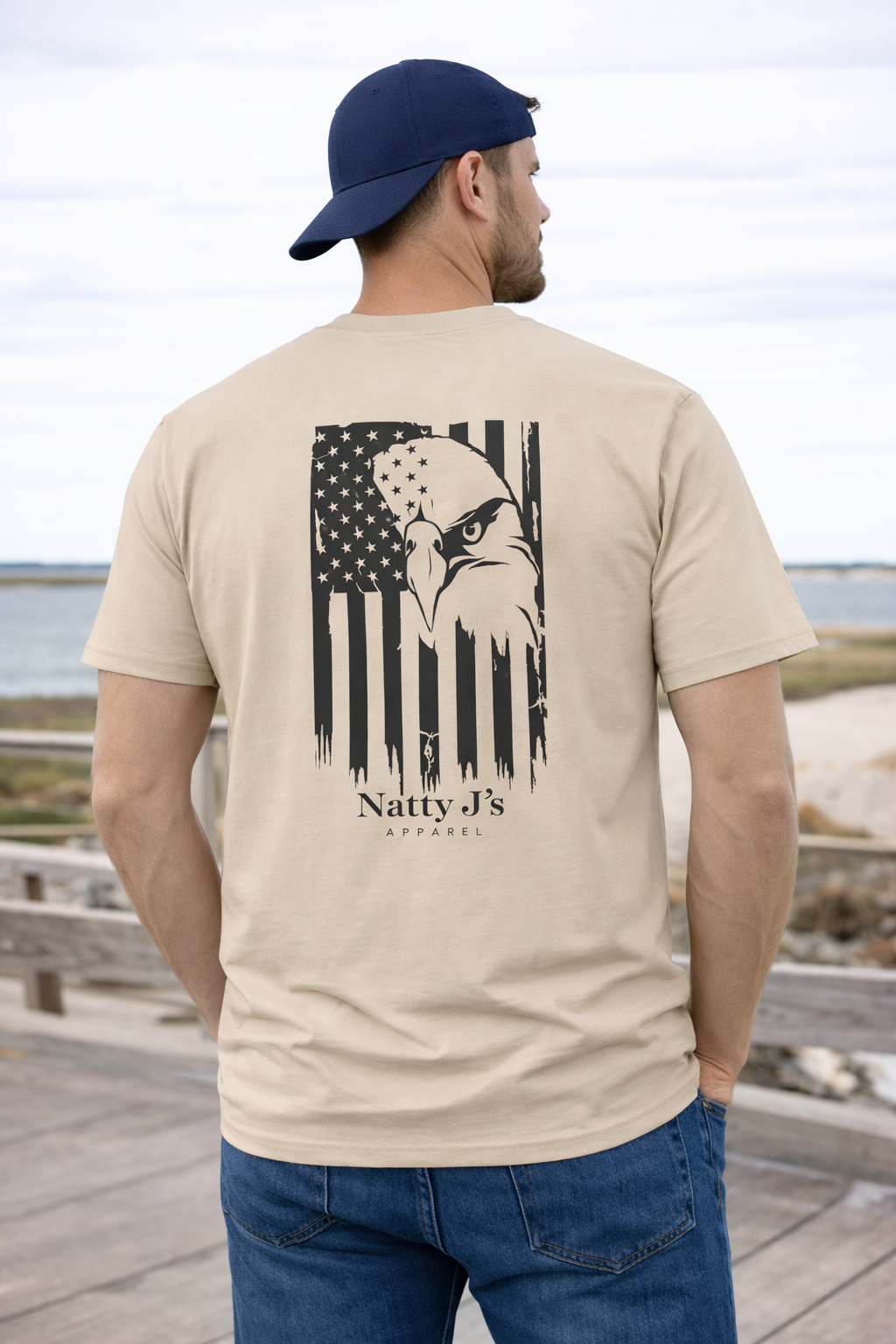 Man wearing a beige t-shirt with an eagle and American flag design, standing on a wooden deck by the water.
