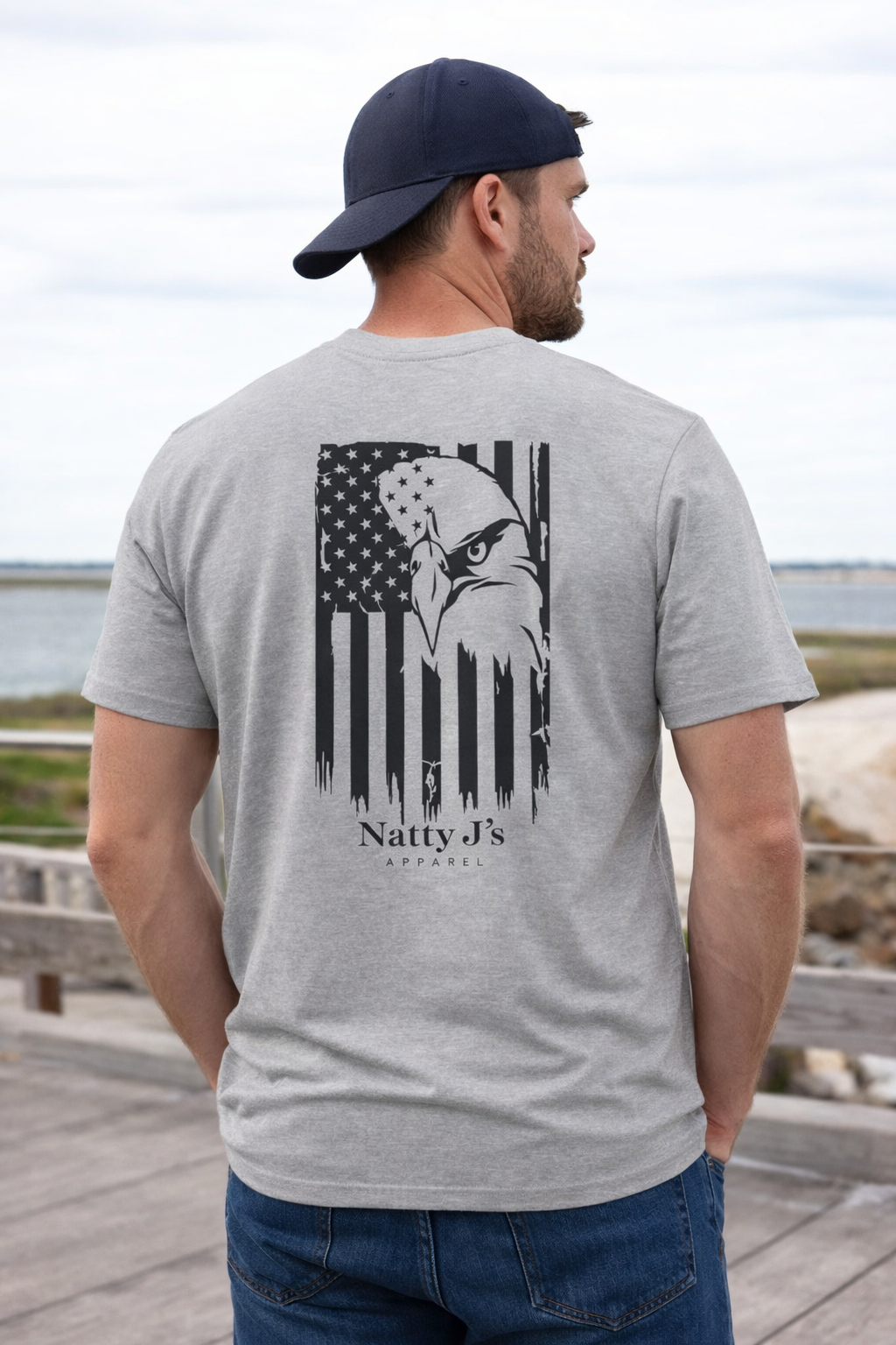 Man wearing a gray t-shirt with an eagle and American flag design, standing on a wooden deck by a body of water.
