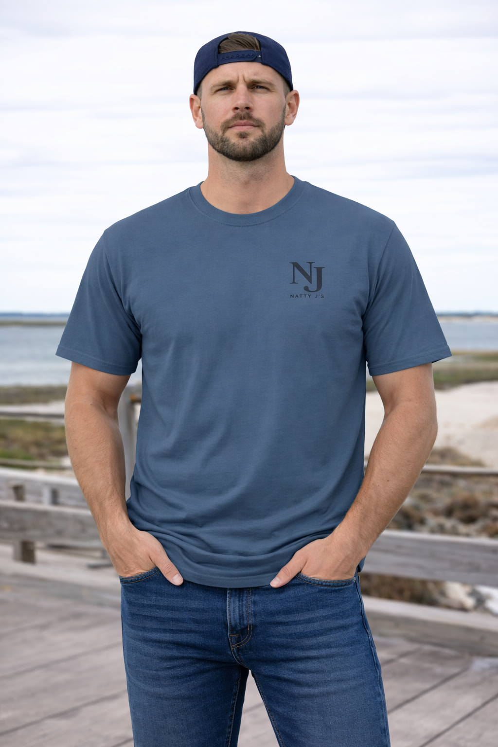 Man wearing a blue t-shirt with a logo and cap, standing on a wooden boardwalk by the beach.
